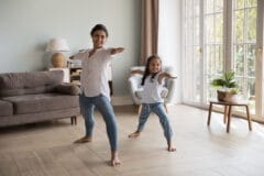 Young Indian woman and little girl perform asanas at modern home. Mom teach daughter yoga do together Warrior one exercise, standing barefoot in cozy warm living room. Healthy lifestyle, sport concept